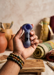 Hand holding a small purple smoking pipe with a blurred background of rustic items.